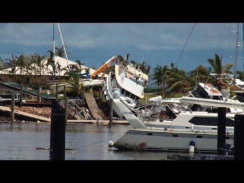 Destroyed and overturned vessels piled against a marina jetty with palm trees visible in the background — the aftermath of Cyclone Larry's storm surge and winds