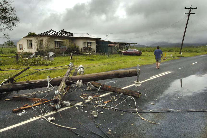 A person walks down a road past a house with its roof blown off. A big power pole is broken and lying across the wet road after Cyclone Larry