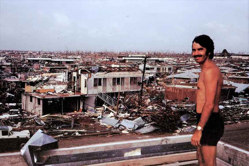 A man who lives in Darwin is standing on a roof. He is looking at the broken houses below. The photo was taken in 1974.